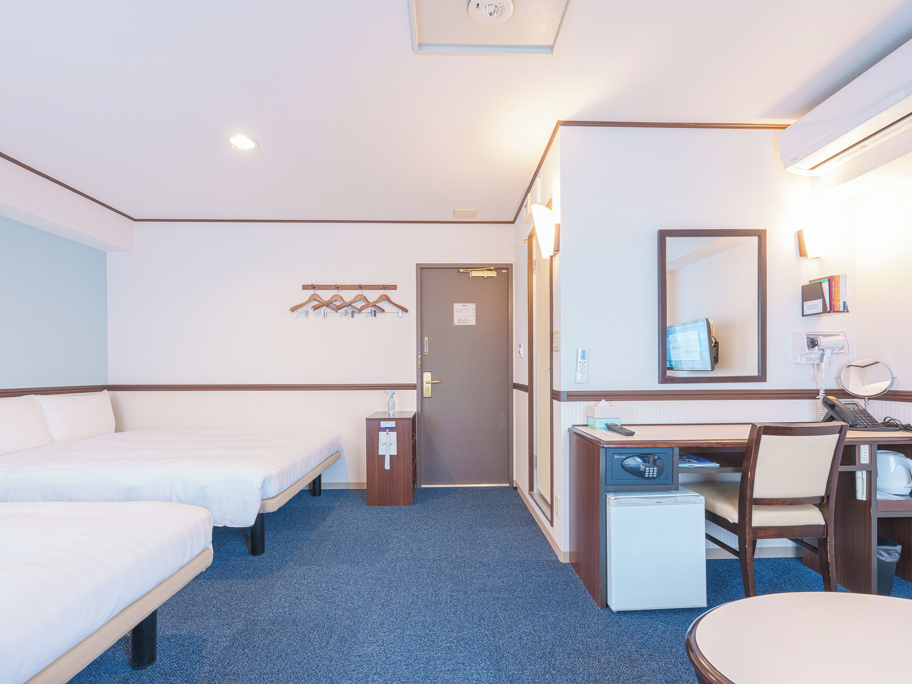 A hotel room with two beds, a desk, a chair, a small refrigerator, a mirror, and a wall-mounted TV.