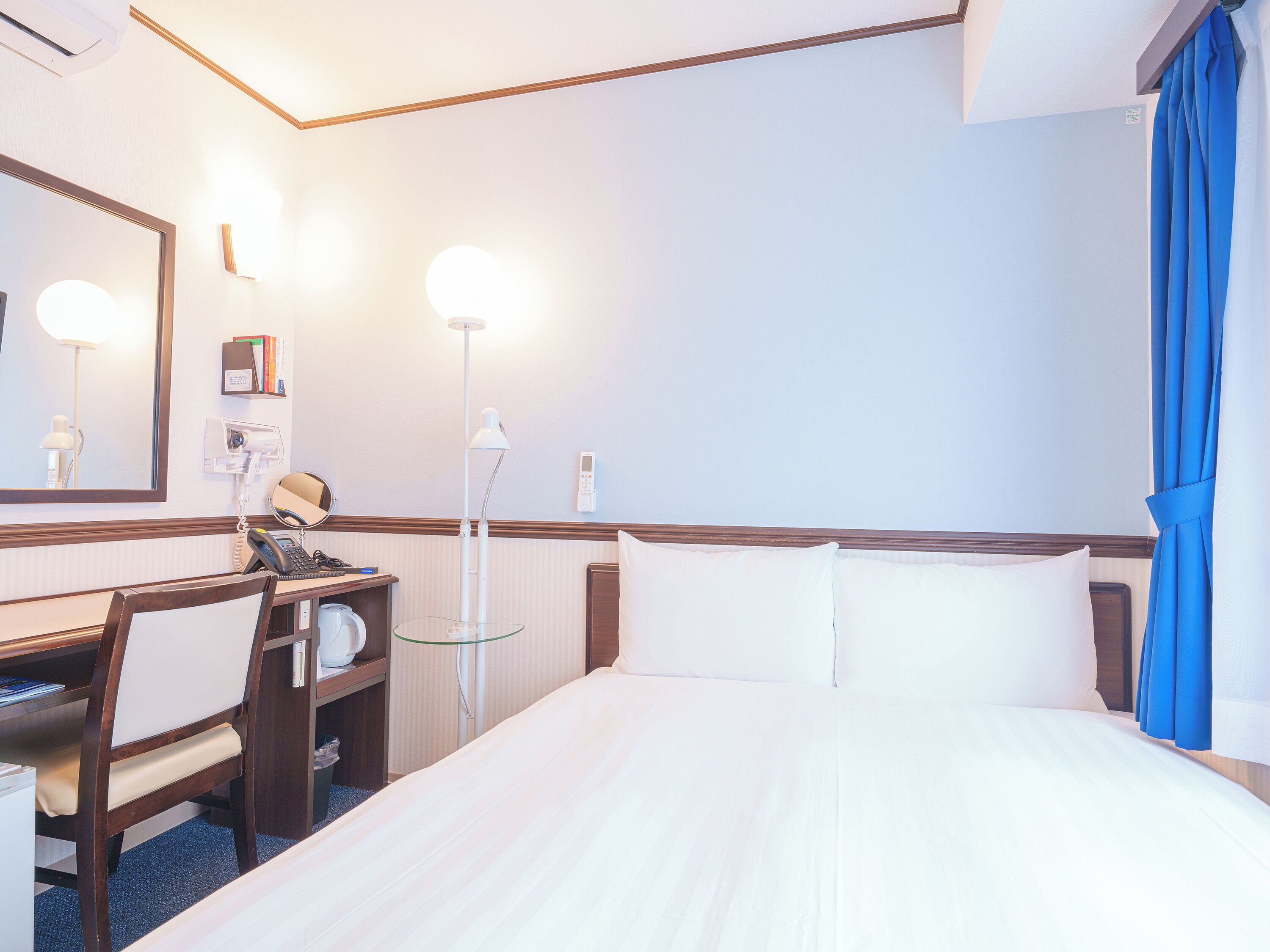 A hotel room with a bed, a desk with a telephone, a mirror, and a lamp.