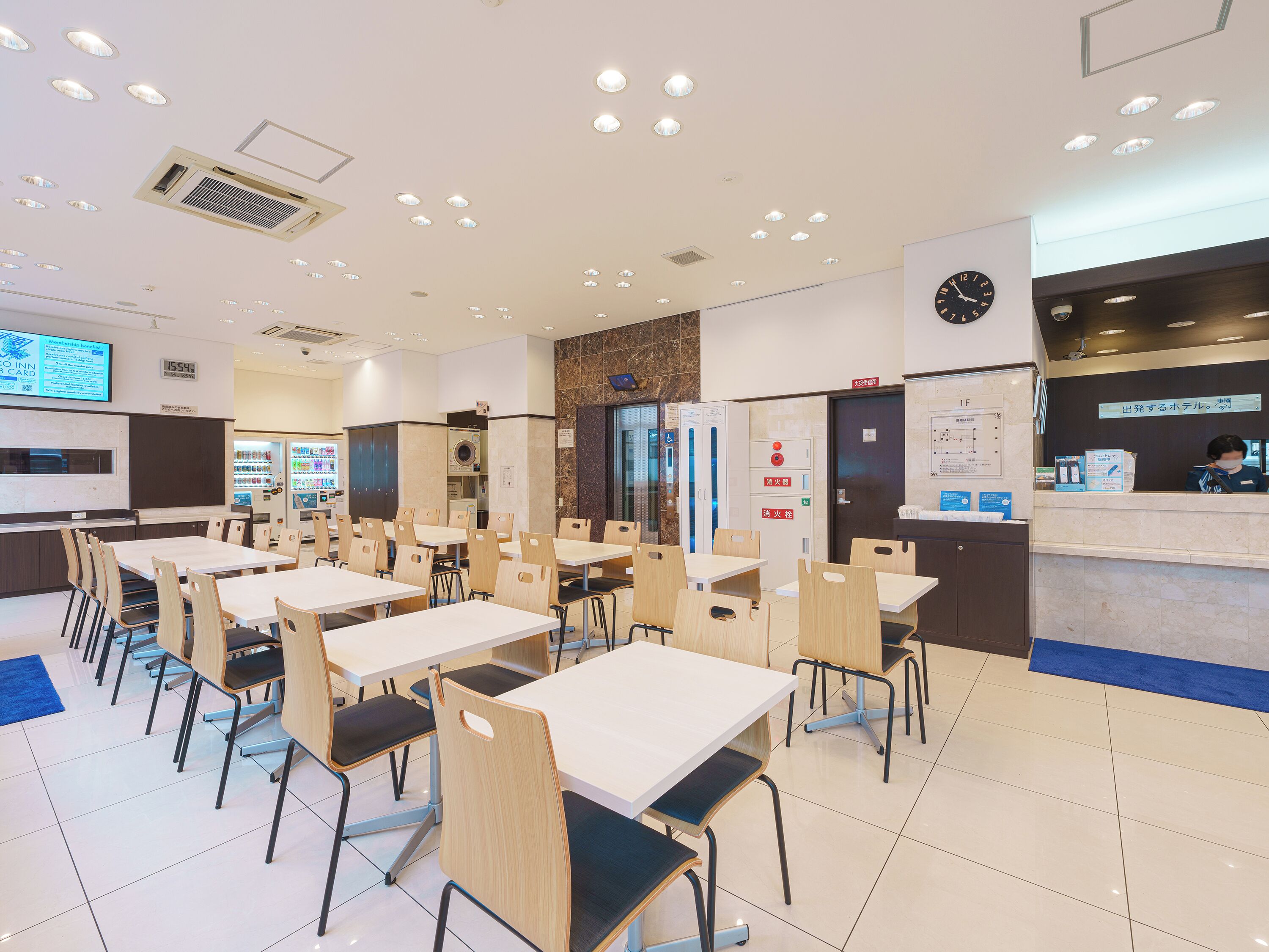 Comedor de hotel con varias mesas y sillas, una recepción y un reloj en la pared.