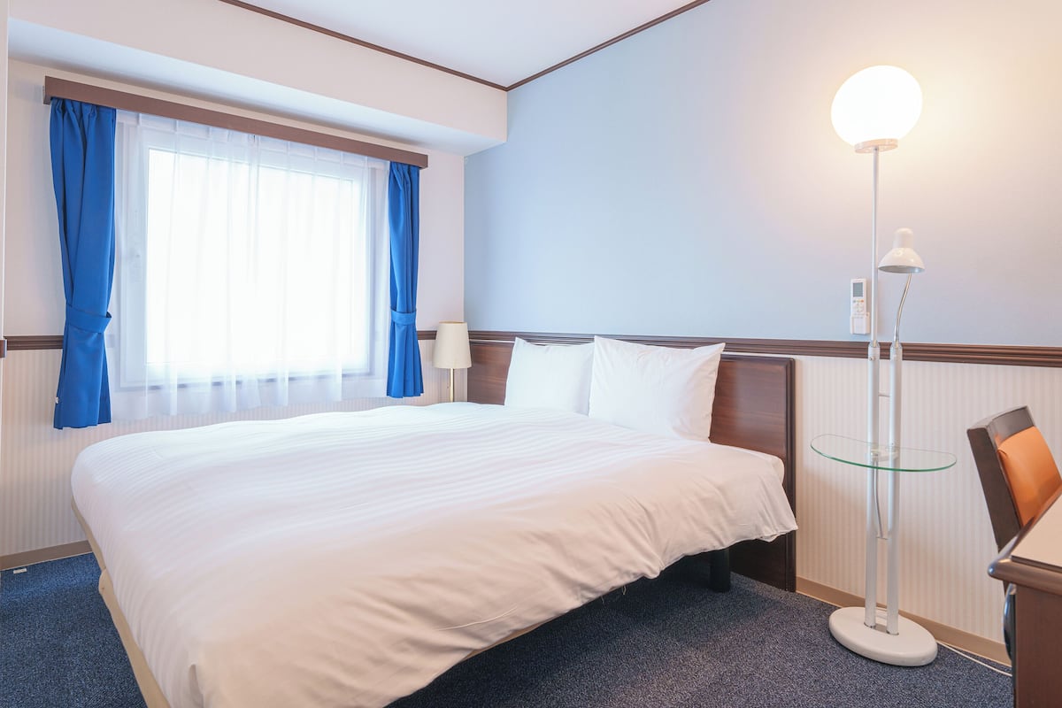 Une chambre d’hôtel avec un grand lit, une table de chevet avec une lampe, une fenêtre avec des rideaux bleus et un panneau de commande fixé au mur.