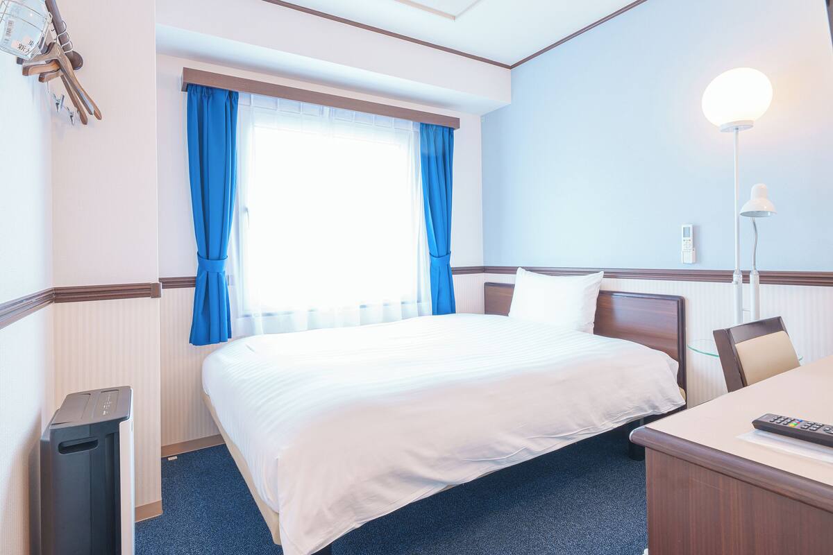 Une chambre d’hôtel avec un lit, un bureau, une lampe et une fenêtre avec des rideaux.