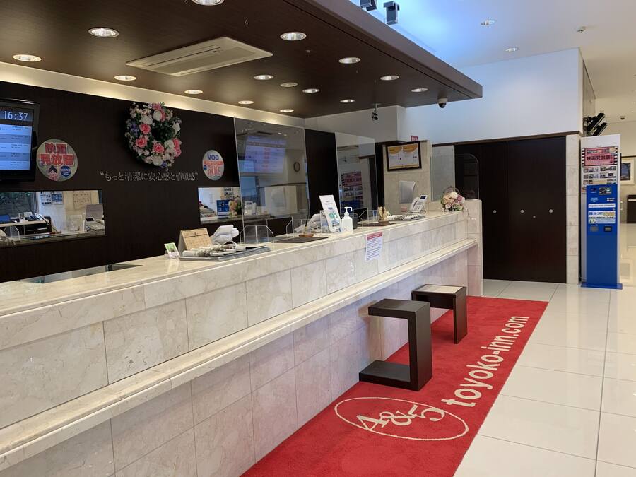 A hotel reception desk with a digital clock, a floral decoration, and a red carpet with a logo.