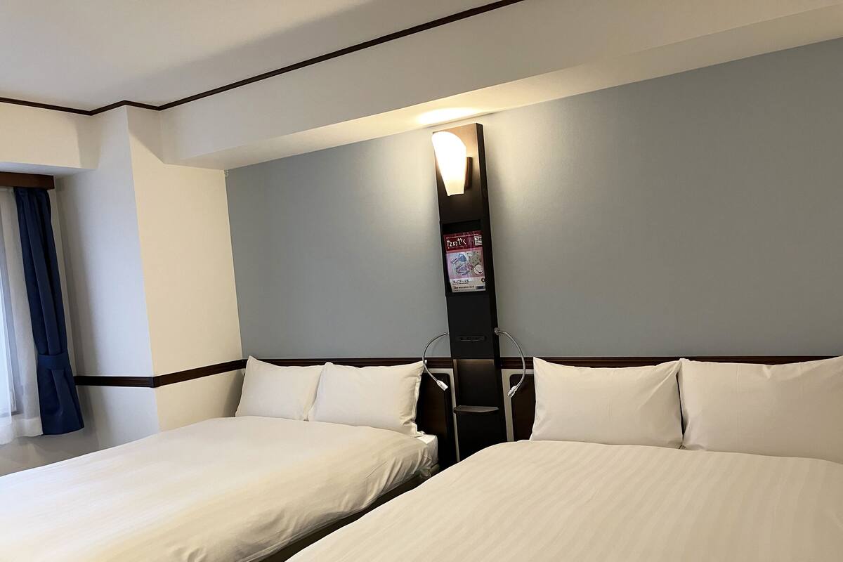 Habitación de hotel con dos camas, una mesita de noche, una lámpara de pared y una ventana con cortinas.