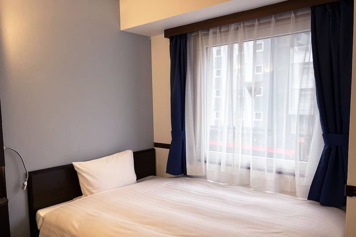 Una habitación de hotel con una cama, una ventana con cortinas translúcidas y vistas a edificios.