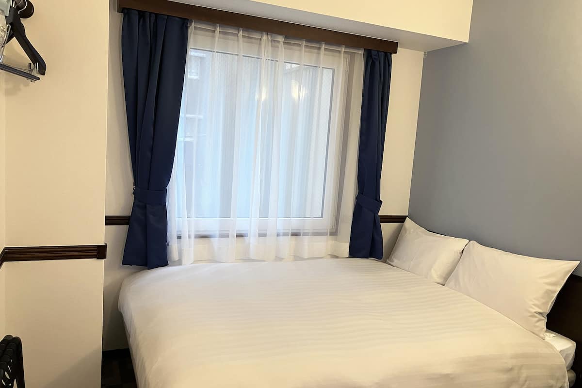 Una cama bien hecha en una habitación con una ventana cubierta por cortinas blancas translúcidas y cortinas azul oscuro.