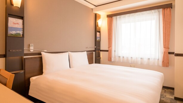 Une chambre d’hôtel avec un grand lit, un bureau, une chaise, une table de chevet, une lampe fixée au mur et une fenêtre avec des rideaux.