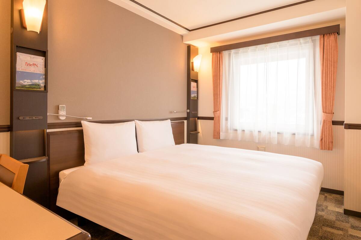 Une chambre d’hôtel avec un grand lit, un bureau, une chaise, une table de chevet, une lampe fixée au mur et une fenêtre avec des rideaux.