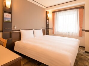 Une chambre d’hôtel avec un grand lit, un bureau, une chaise, une table de chevet, une lampe fixée au mur et une fenêtre avec des rideaux.