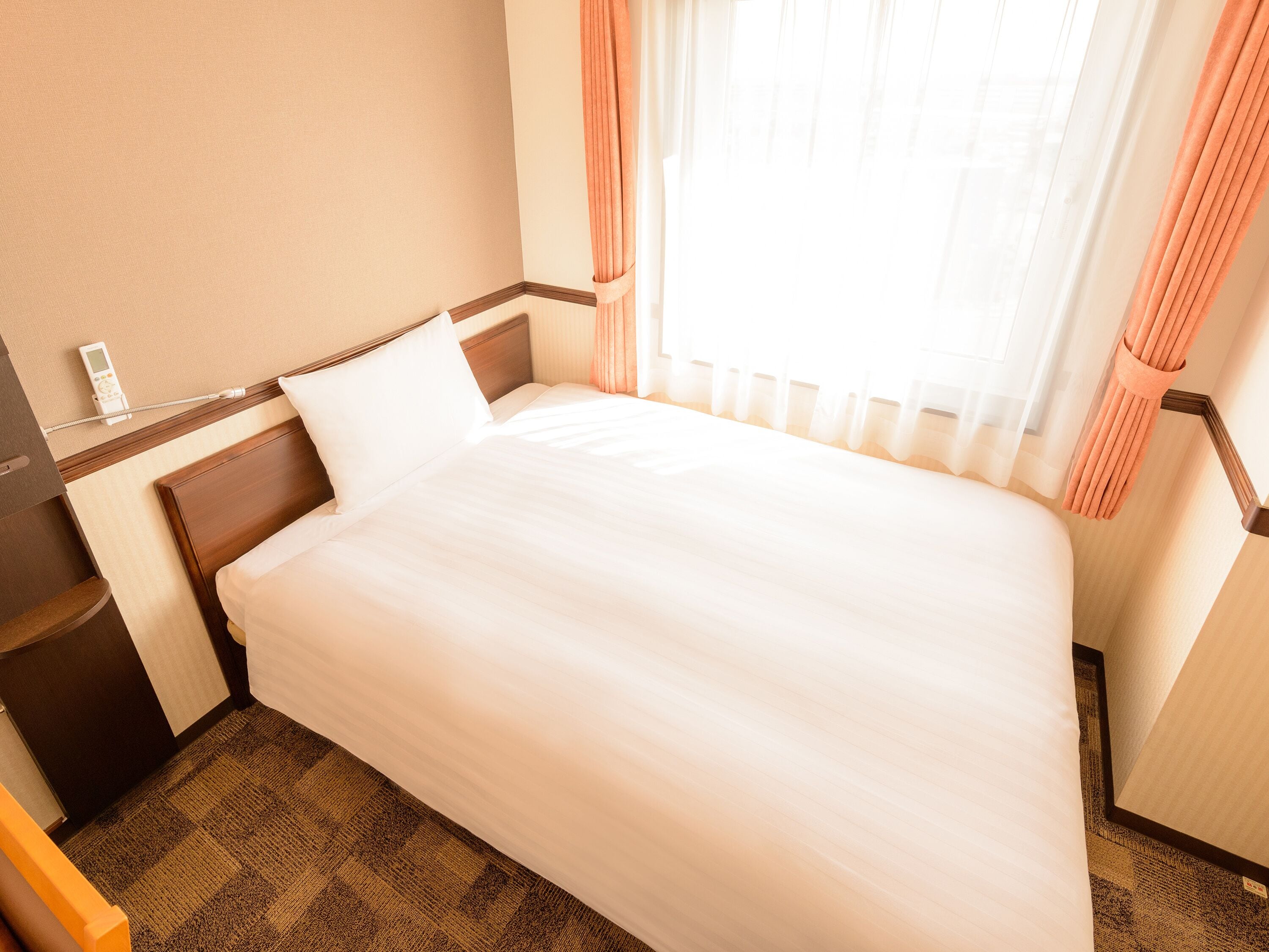 A neatly made bed in a hotel room with a wooden headboard, a bedside table with a lamp, and a window with sheer curtains.