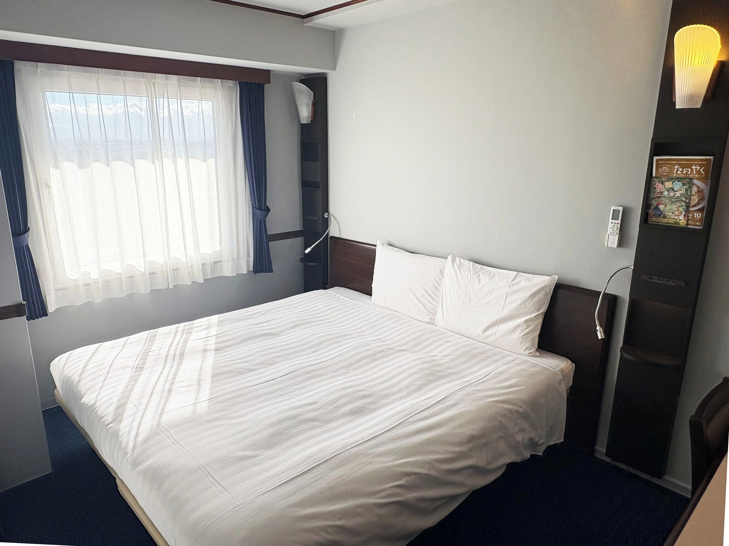 A hotel room with a bed, a window with curtains, a bedside table, and a wall-mounted light fixture.