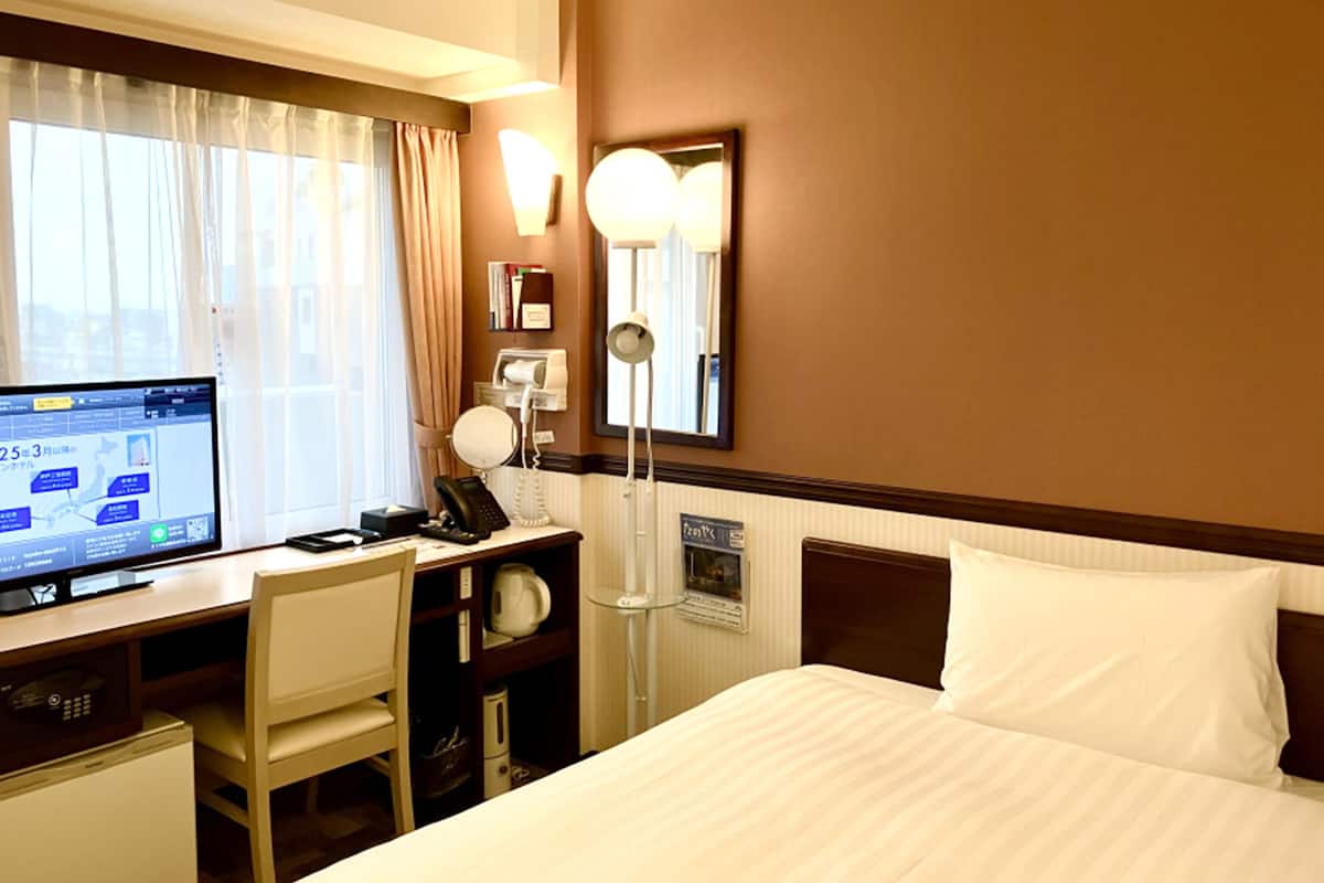 Une chambre d’hôtel comprenant un lit, un bureau avec un ordinateur, une chaise, un téléphone, un miroir et une lampe fixée au mur.