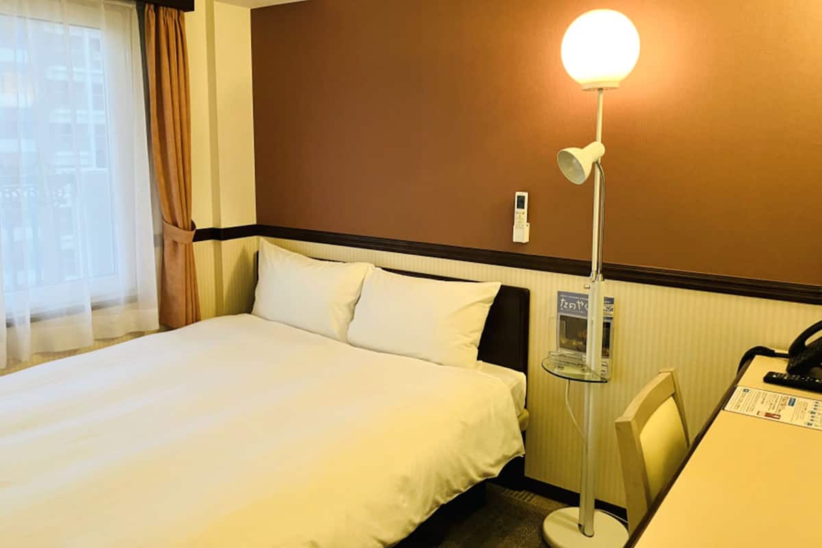 Une chambre d’hôtel avec un lit, un bureau, une lampe et un thermostat fixé au mur.