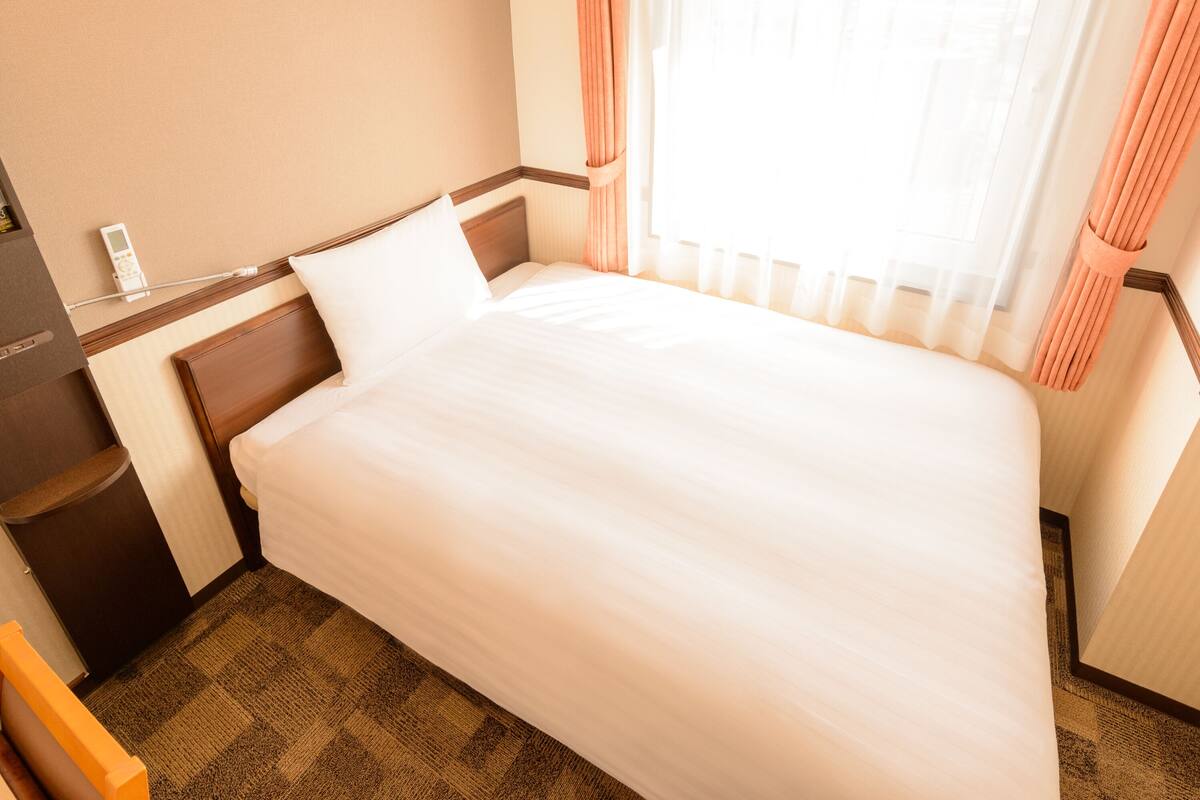 Ein Hotelzimmer mit einem Einzelbett, einem Nachttisch mit Lampe, einem kleinen Beistelltisch und einem Fenster mit Vorhängen.