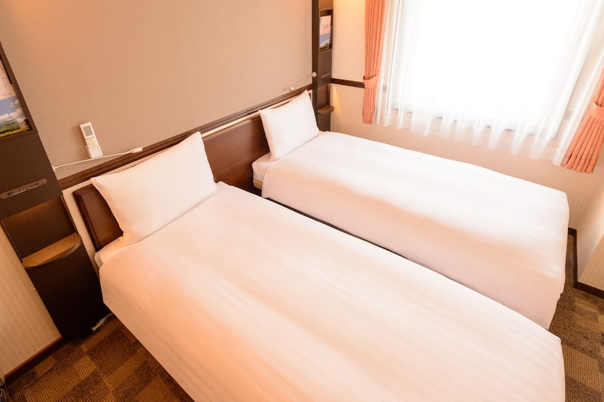 Ein Hotelzimmer mit zwei Einzelbetten, einem Nachttisch, einem Telefon und einem Vorhangfenster.