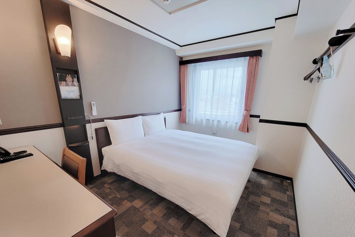 Ein Hotelzimmer mit einem Bett, einem Schreibtisch, einem Sessel, einem Nachttisch, einem Fenster mit Vorhängen und einer Wandleuchte.