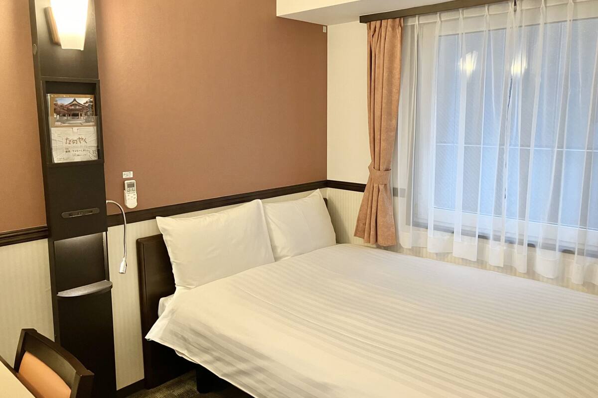 A hotel room with a bed, a desk, a chair, a wall-mounted phone, and a window with sheer curtains.