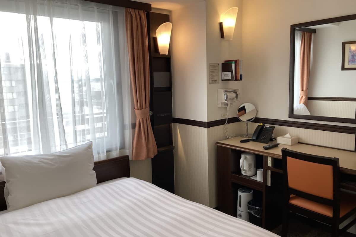 A hotel room with a bed, a desk with a telephone, a mirror, and a window with curtains.
