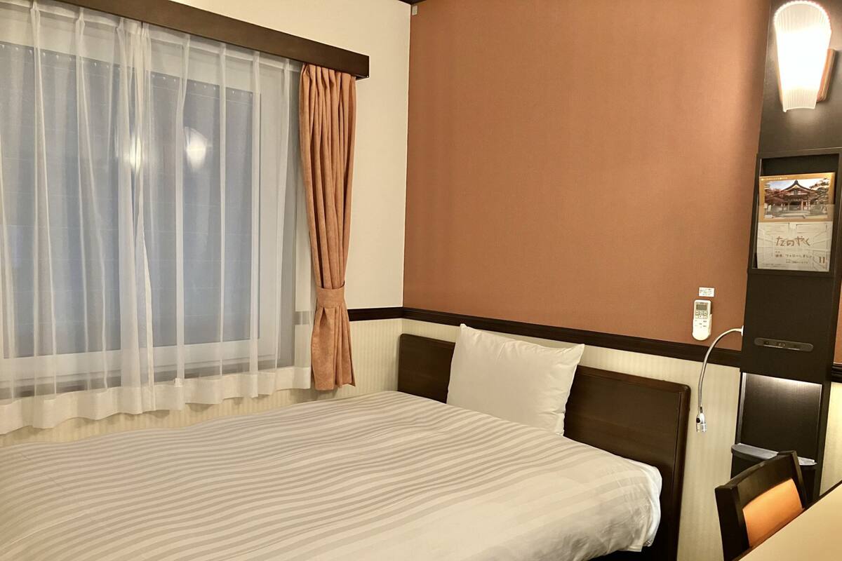A hotel room with a large bed, a bedside table, a lamp, and a window with sheer curtains and a tieback.