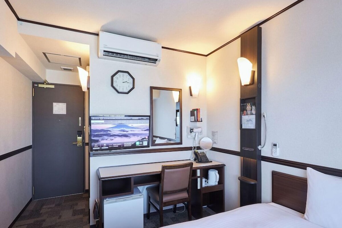 Habitación de hotel con cama, escritorio con silla, reloj, espejo, refrigerador pequeño y aire acondicionado de pared.