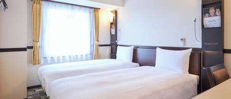 Quarto de hotel com duas camas, uma janela grande com cortinas, uma luminária na parede e uma pequena escrivaninha.