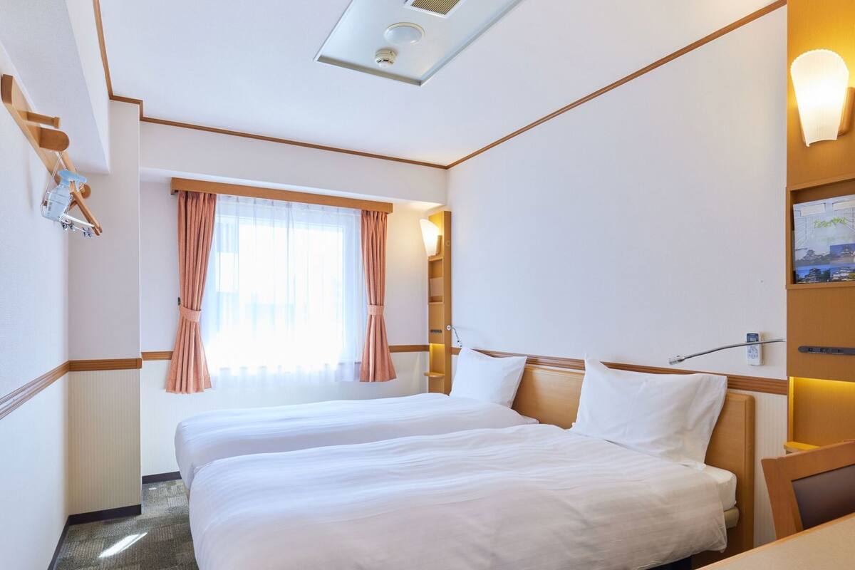 A hotel room with two beds, a window with curtains, a wall-mounted lamp, and a ceiling vent.