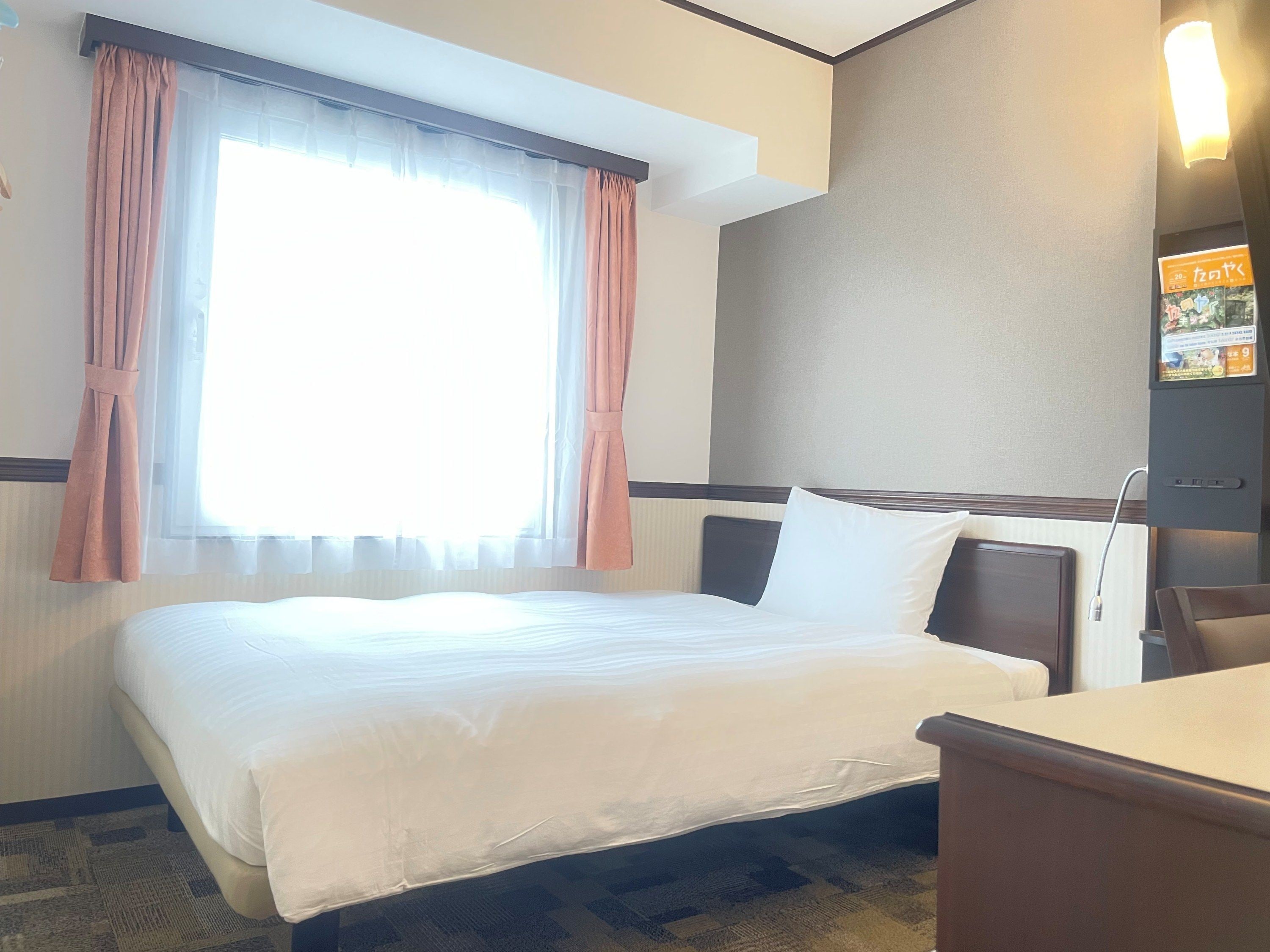 A hotel room with a bed, a desk, a window with curtains, and a wall-mounted TV.
