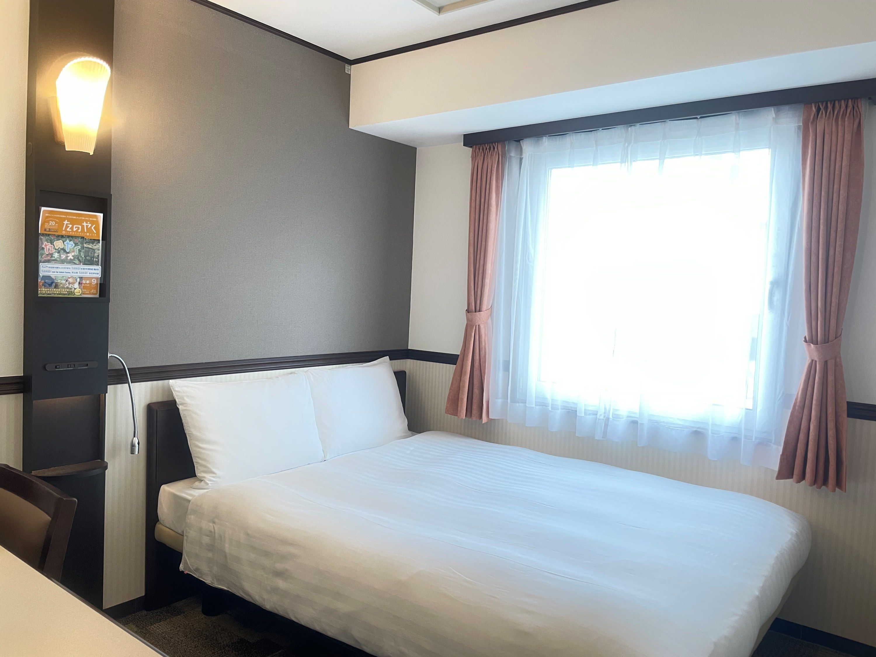 A hotel room with a bed, a desk, a chair, a window with curtains, and a wall-mounted light fixture.