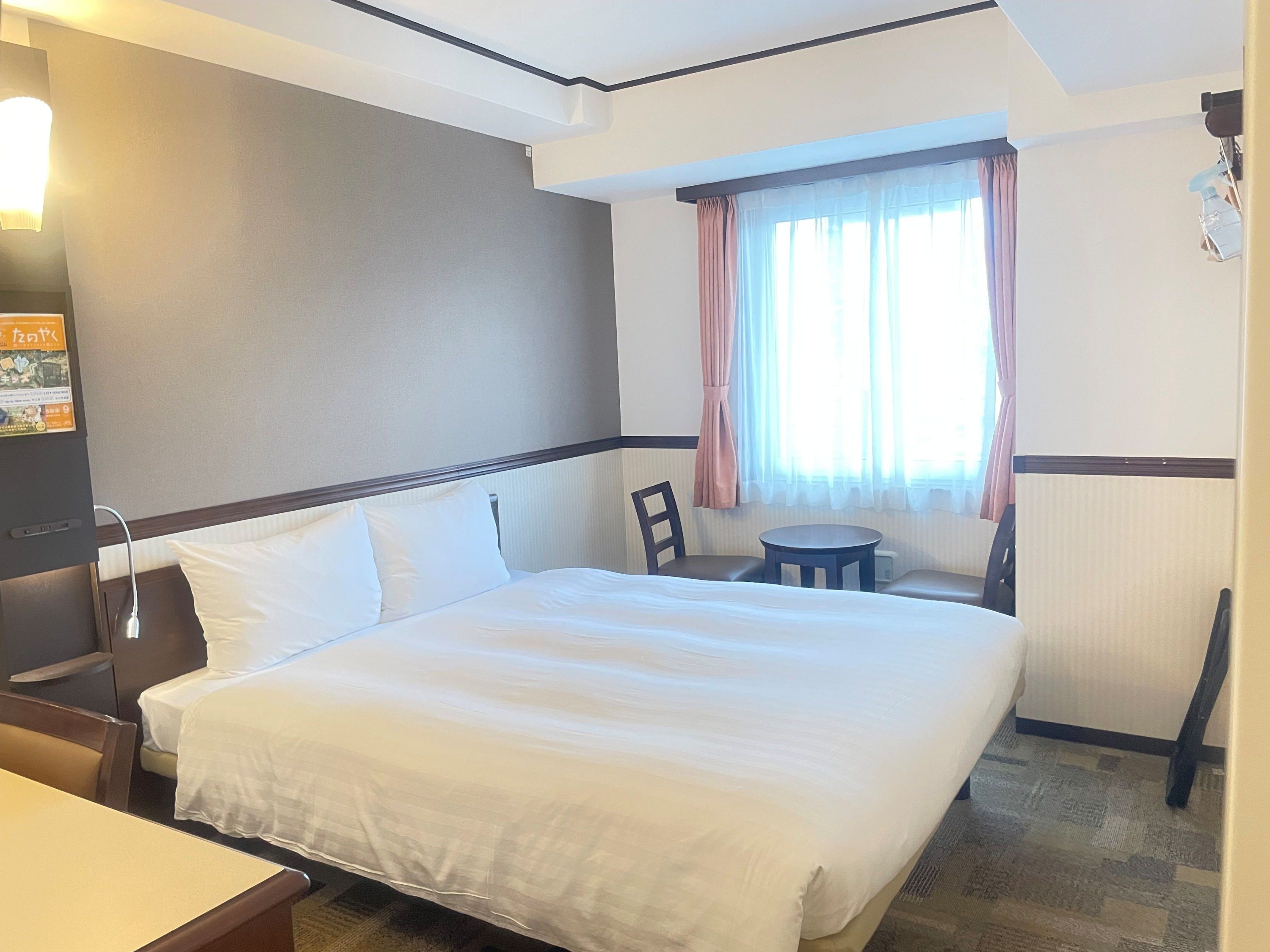 A hotel room with a bed, a small table, a chair, and a window with curtains.