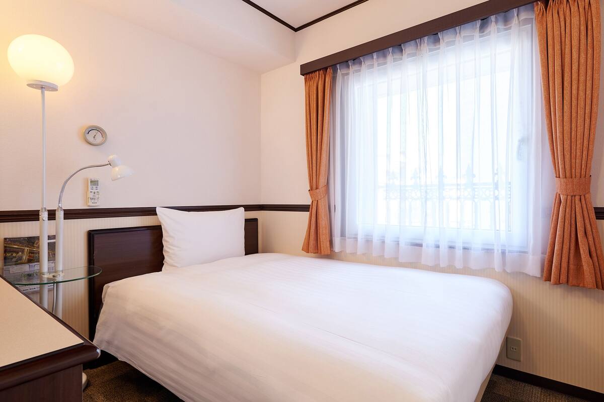 A hotel room with a neatly made bed, a bedside lamp, a clock on the wall, and a window with curtains.