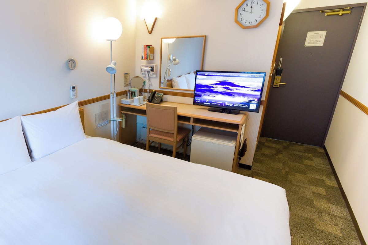A hotel room with a bed, desk, chair, computer, and a clock on the wall.