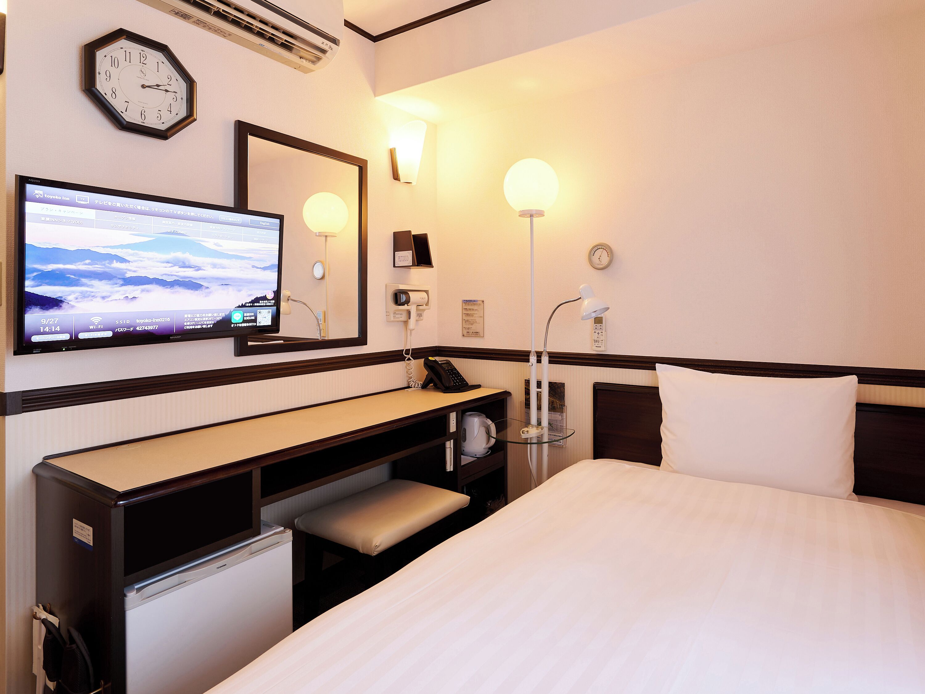 A hotel room with a bed, a desk with a chair, a television, a clock, and a lamp.