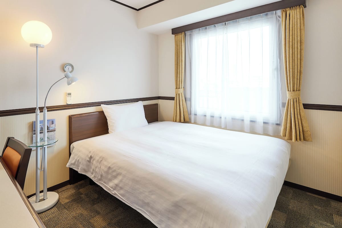 A hotel room with a neatly made bed, a bedside lamp, a clock on the wall, and a window with curtains.