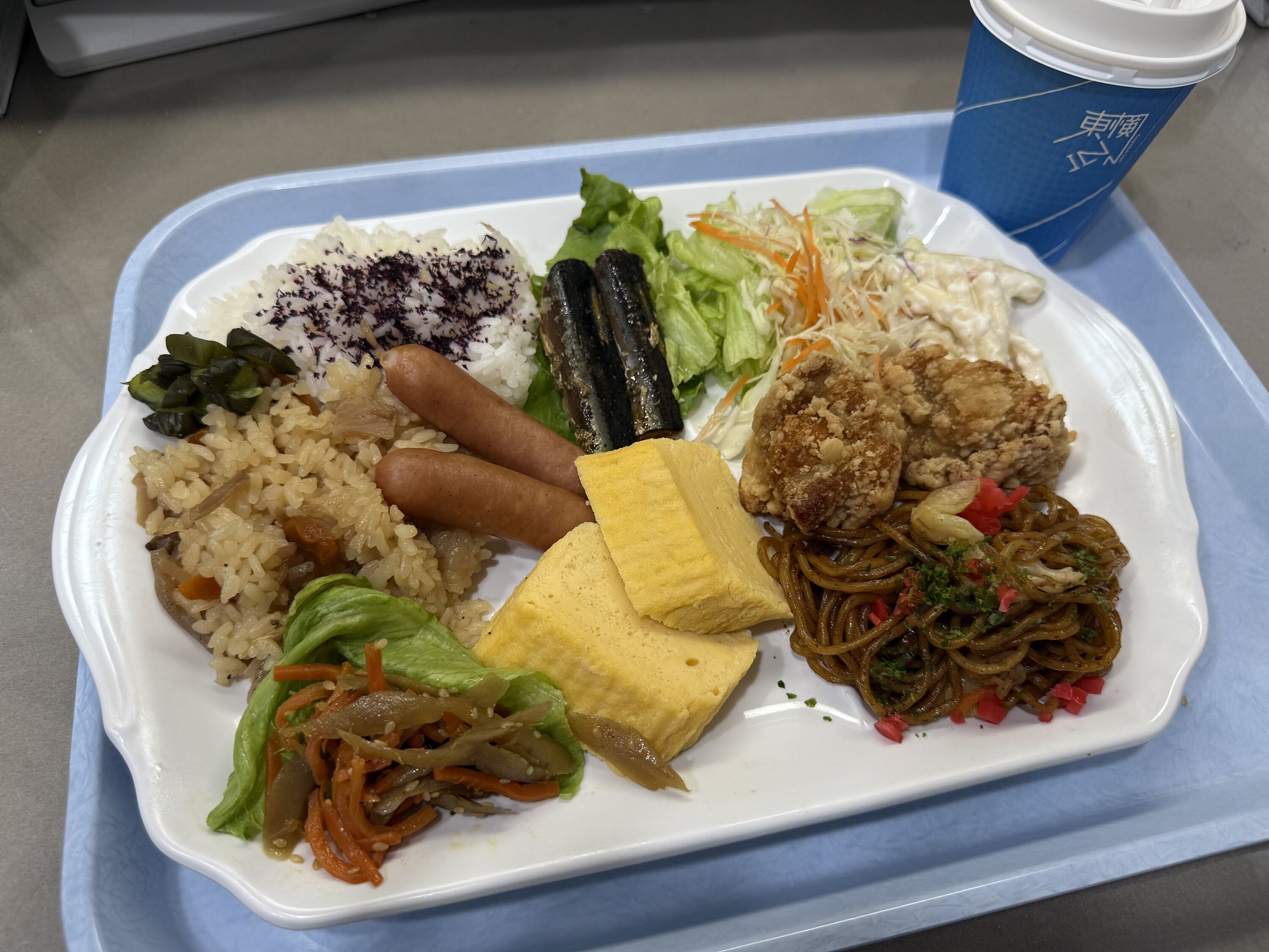 A meal tray with rice, sausages, a fried item, noodles, and a cup.