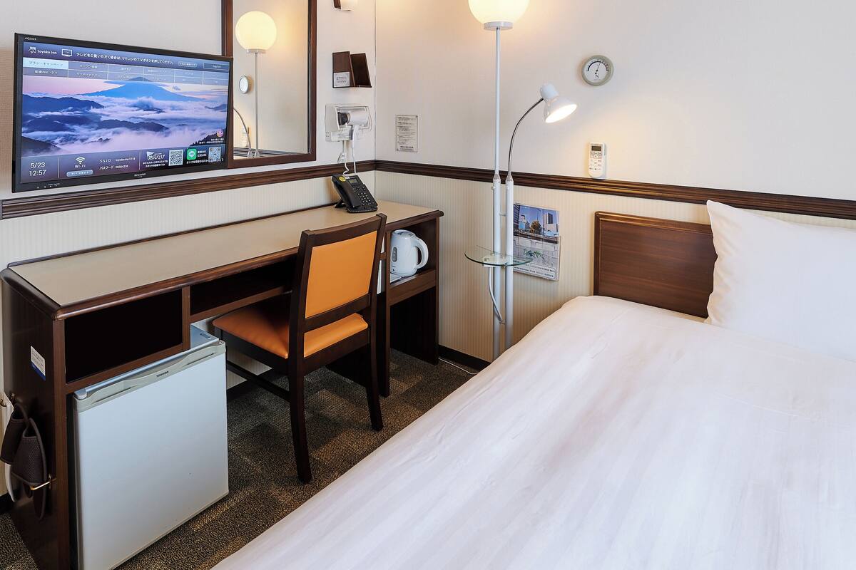 Habitación de hotel con cama, escritorio, silla, un refrigerador pequeño, lámpara de escritorio, reloj y televisor mostrando una imagen panorámica.