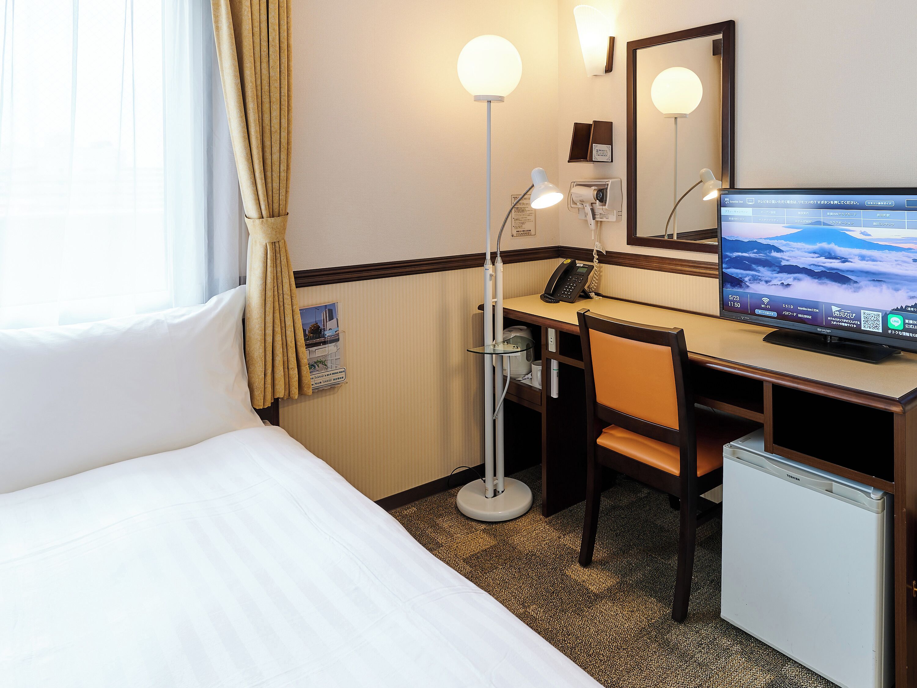 A hotel room with a bed, desk, chair, desk lamp, and a television.