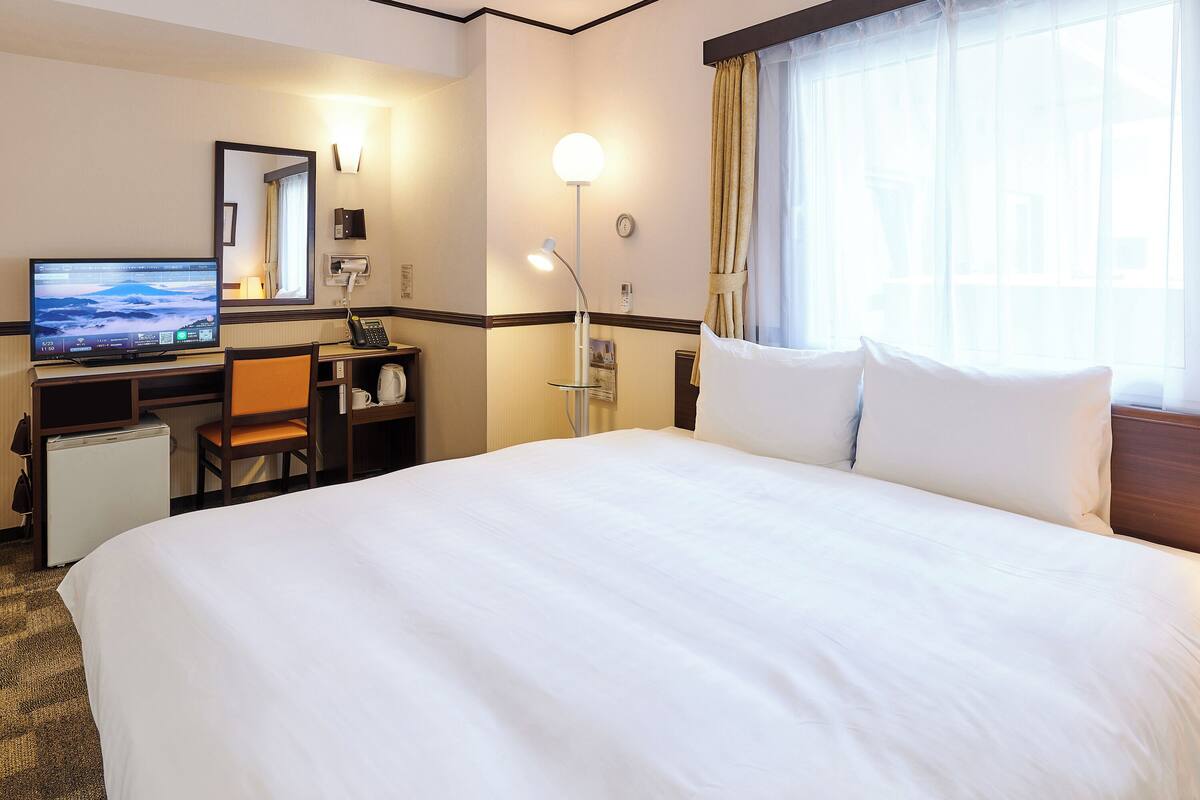 Una habitación de hotel con cama, escritorio, silla, televisor y ventana con cortinas.