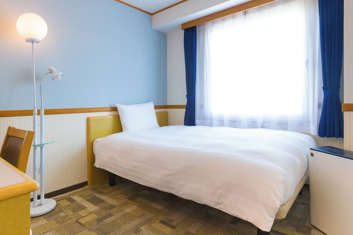 Une chambre d’hôtel avec un lit, un bureau, une lampe et une fenêtre avec des rideaux.