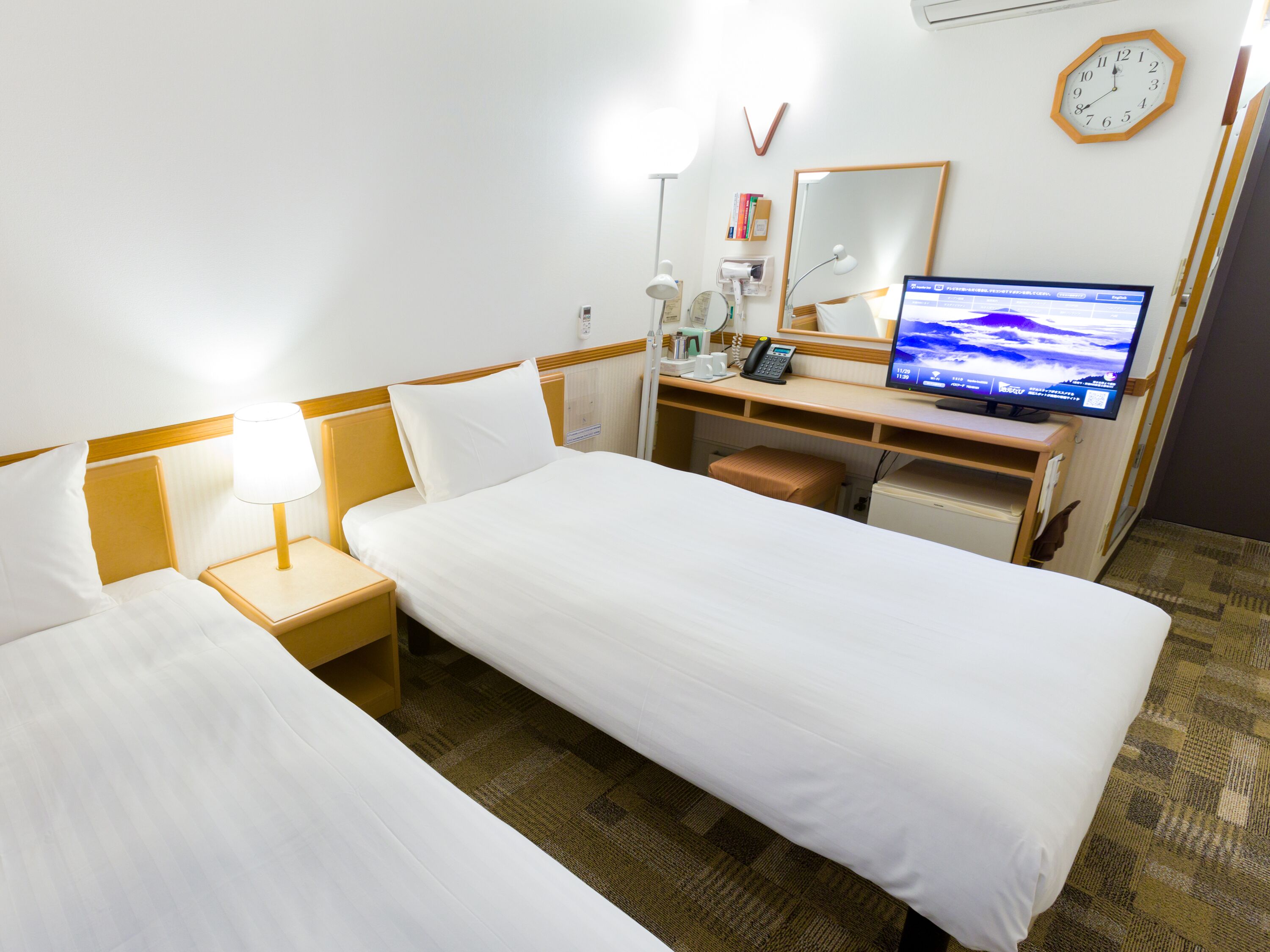 A hotel room with two single beds, a desk with a computer, a clock, and a mirror.