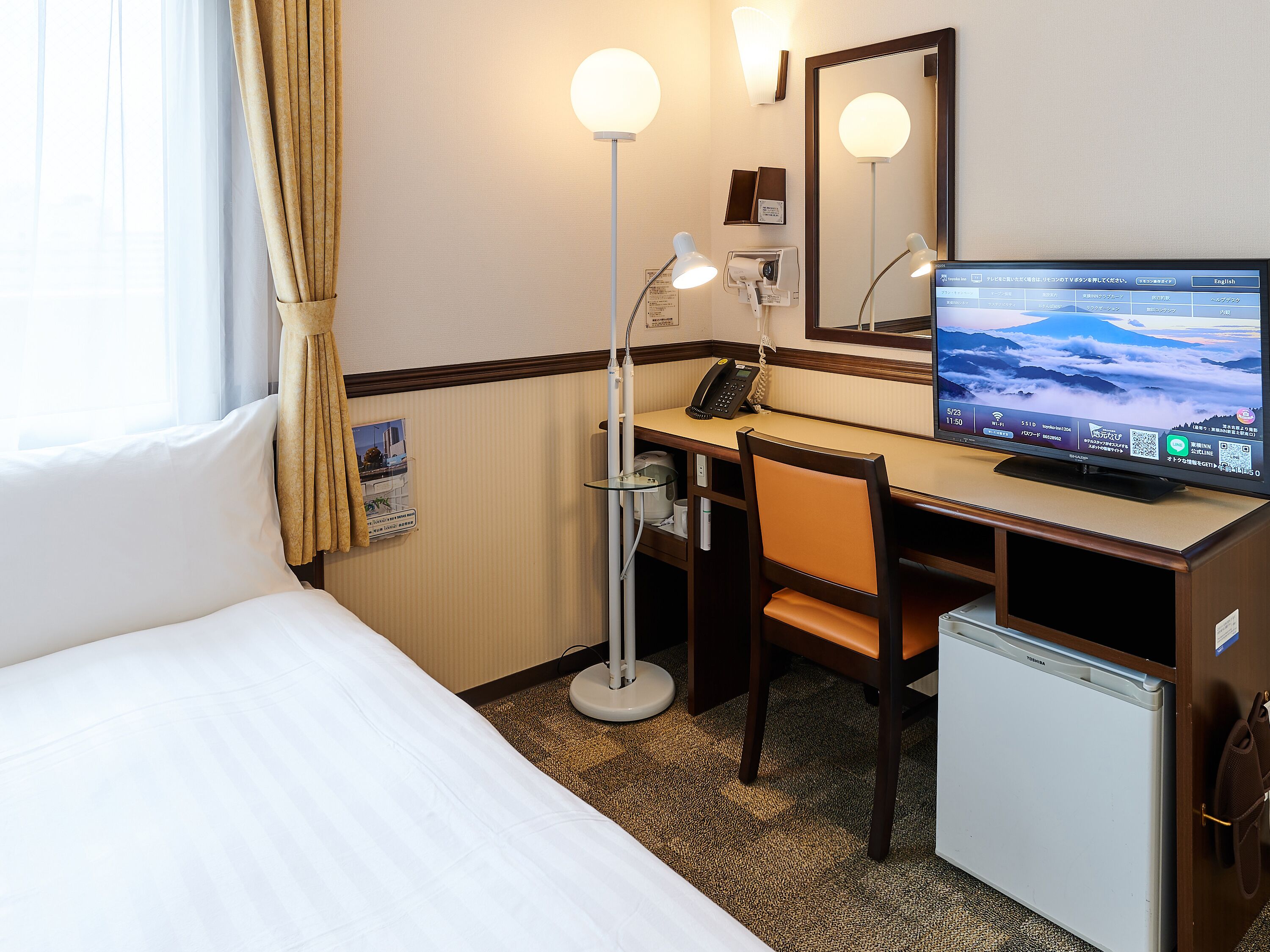 Quarto de hotel com cama, escrivaninha, cadeira, luminária de mesa e televisão.