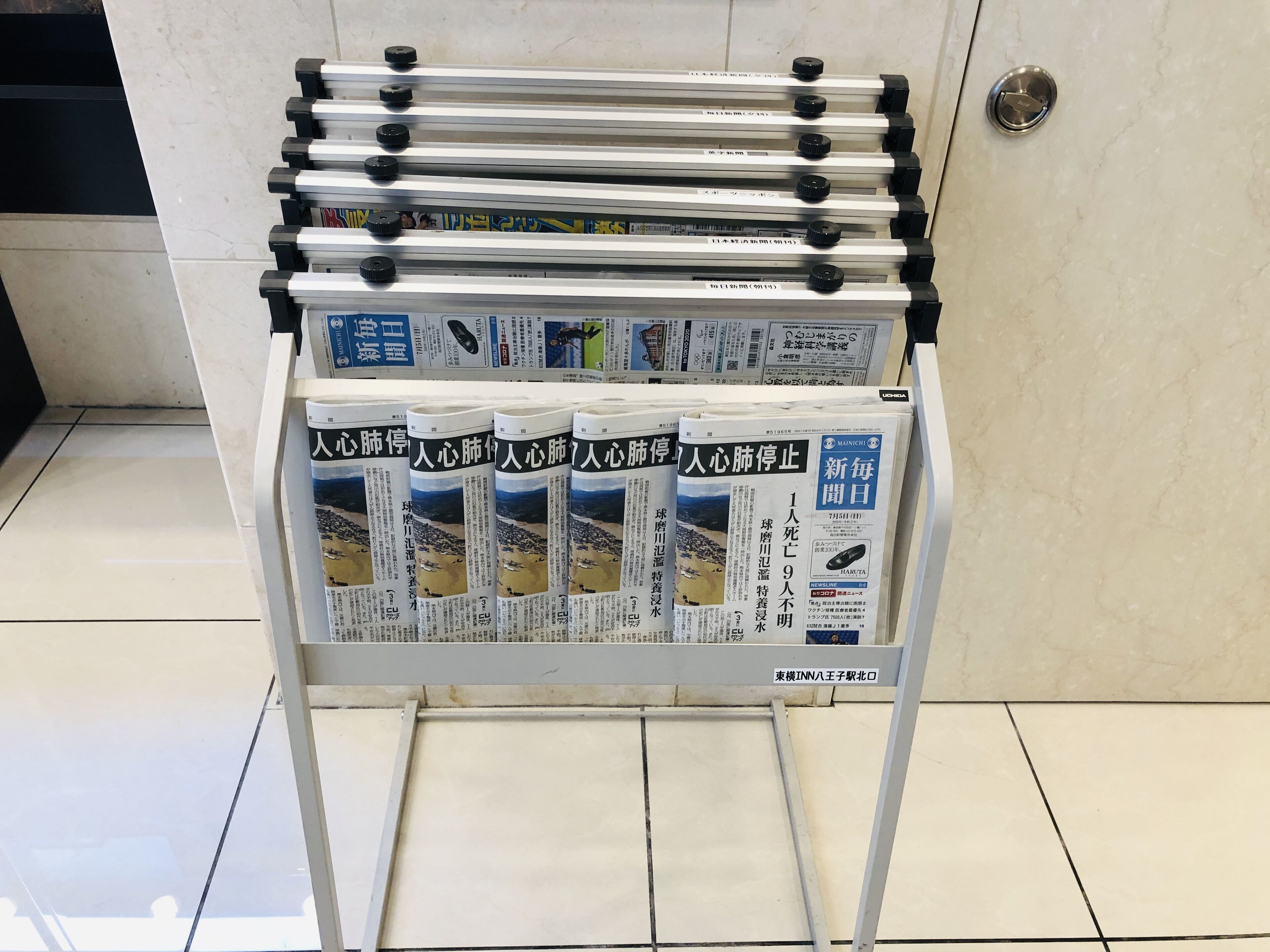 A newsstand with multiple newspapers and magazines, a metal rack with black knobs, and a tiled floor.