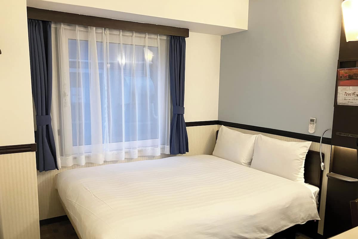 Habitación de hotel con cama, ventana con cortinas translúcidas, mesita de noche y teléfono en la pared.