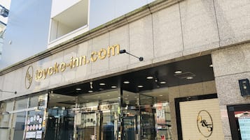 A hotel entrance with a glass facade, a sign reading "toyoko-inn.com", and a digital screen displaying hotel information.