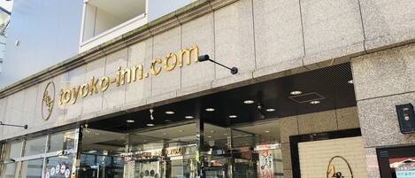 A hotel entrance with a glass facade, a sign reading "toyoko-inn.com", and a digital screen displaying hotel information.