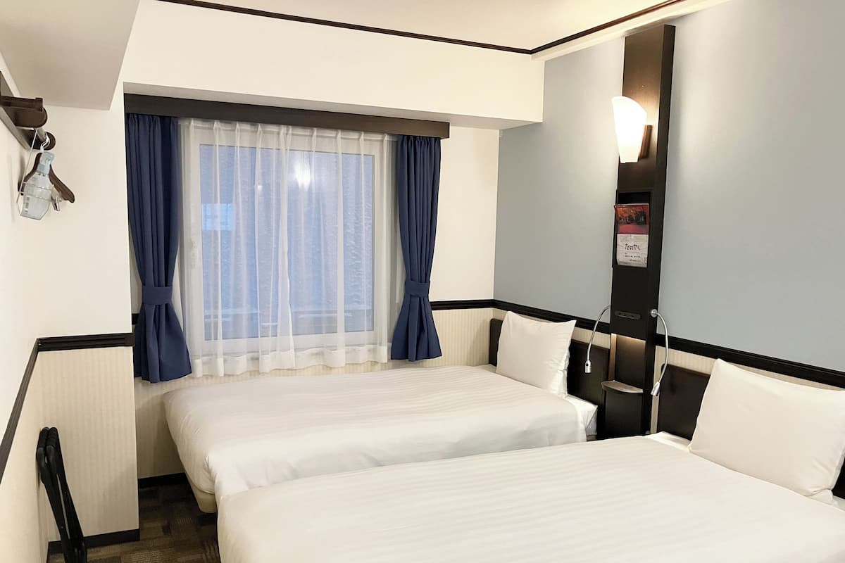 Habitación de hotel con dos camas, una ventana con cortinas translúcidas, una mesita de noche y una lámpara de pared.
