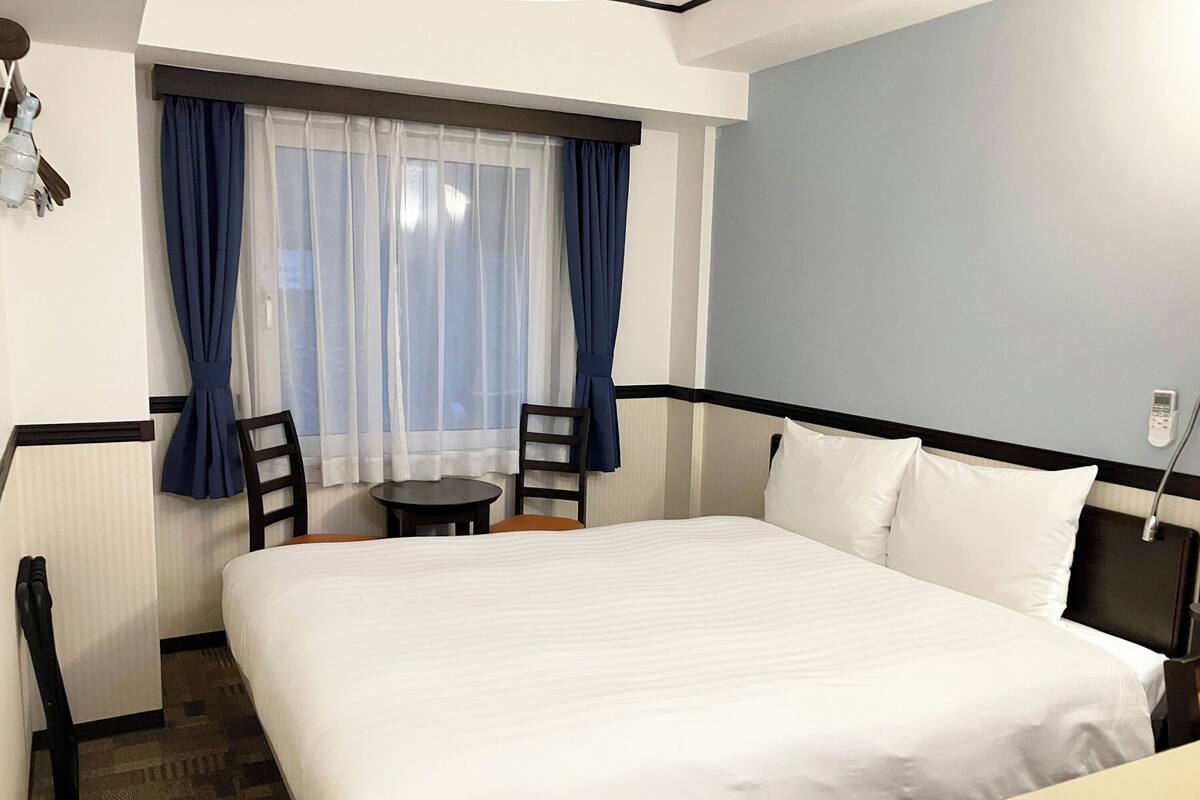 Habitación de hotel con una cama grande, una mesita pequeña, dos sillas y una ventana con cortinas.
