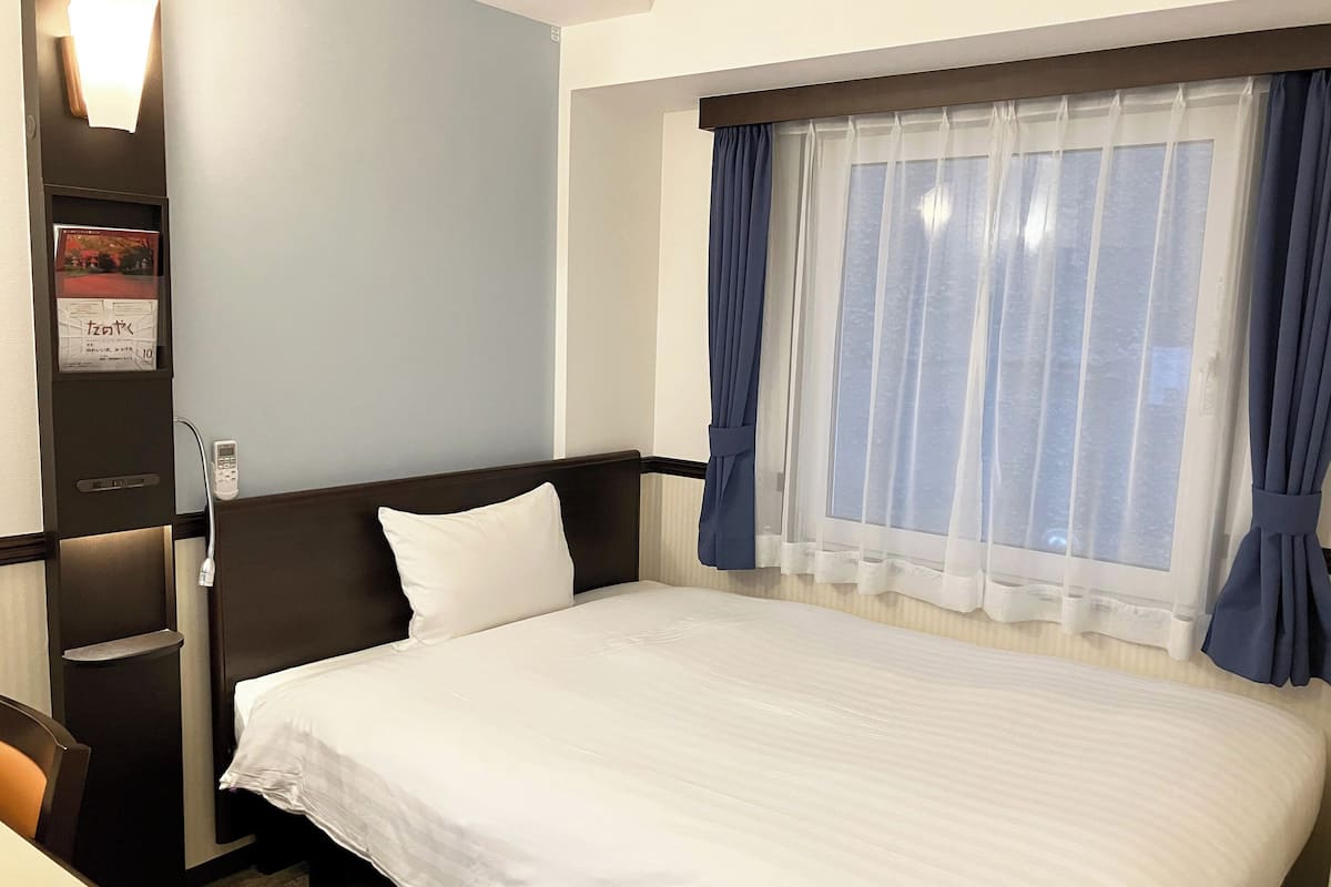 Habitación de hotel con una cama grande, una mesita de noche, una silla, una ventana con cortinas y una lámpara fijada a la pared.