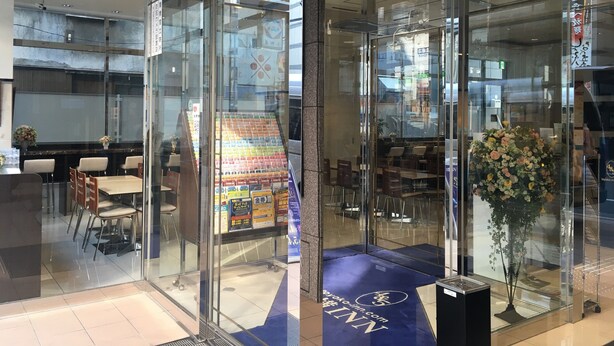 A modern hotel lobby with glass doors, a seating area with tables and chairs, and a display of promotional materials. - Toyoko Inn Tokyo Hachioji Station Kita (Hachioji)