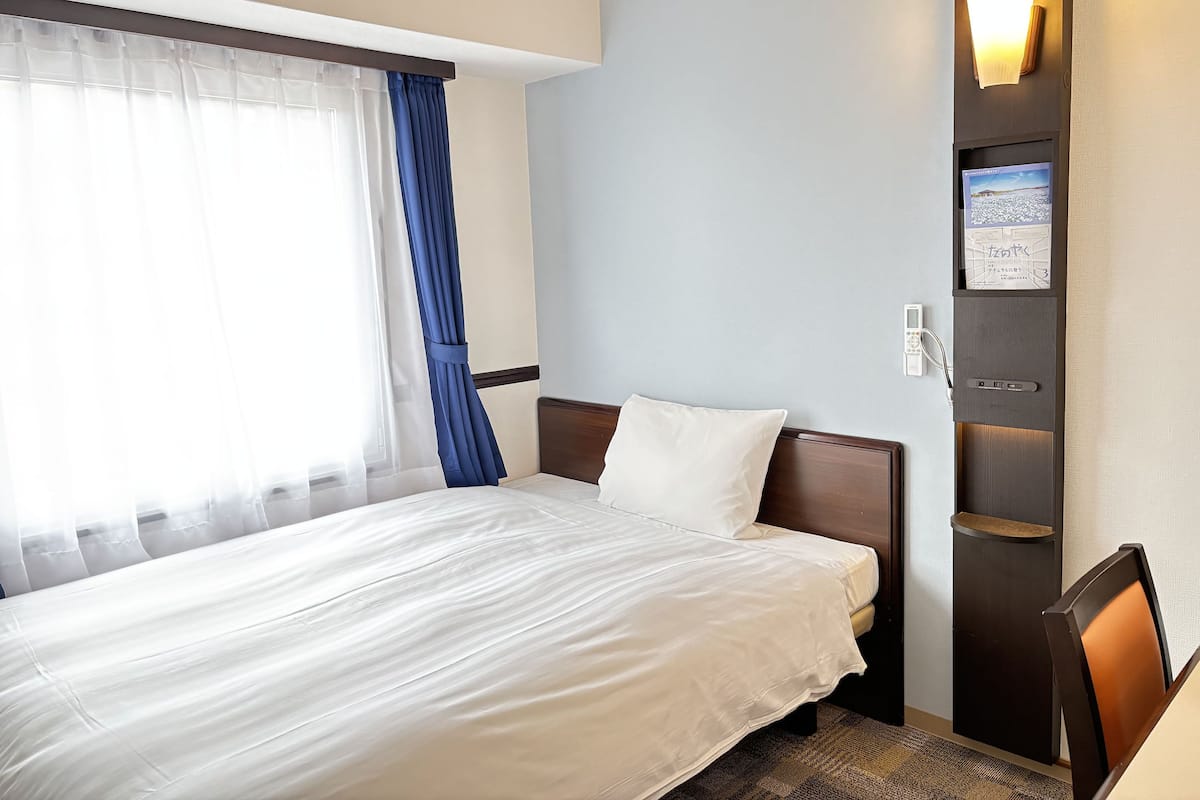 Quarto de hotel com cama, mesa de cabeceira, luminária na parede e janela com cortinas.