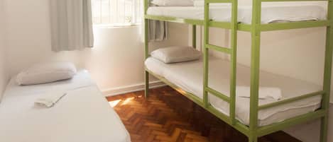 A room with bunk beds and a single bed, wooden parquet flooring, and a window with curtains.
