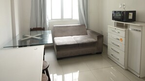 Una habitación pequeña y moderna con una mesa de vidrio, un sofá, un armario blanco con cajones y un microondas encima.